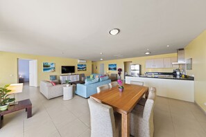A modern living room with a dining table, sofas, and a kitchen area with a refrigerator and stove.