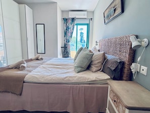 A bedroom with a bed, a bedside table, a lamp, a wall-mounted heater, a window with curtains, and a view of greenery outside.