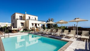 Un espace piscine avec des chaises longues et des parasols, un bâtiment doté d’un balcon, et un ciel d’un bleu limpide.