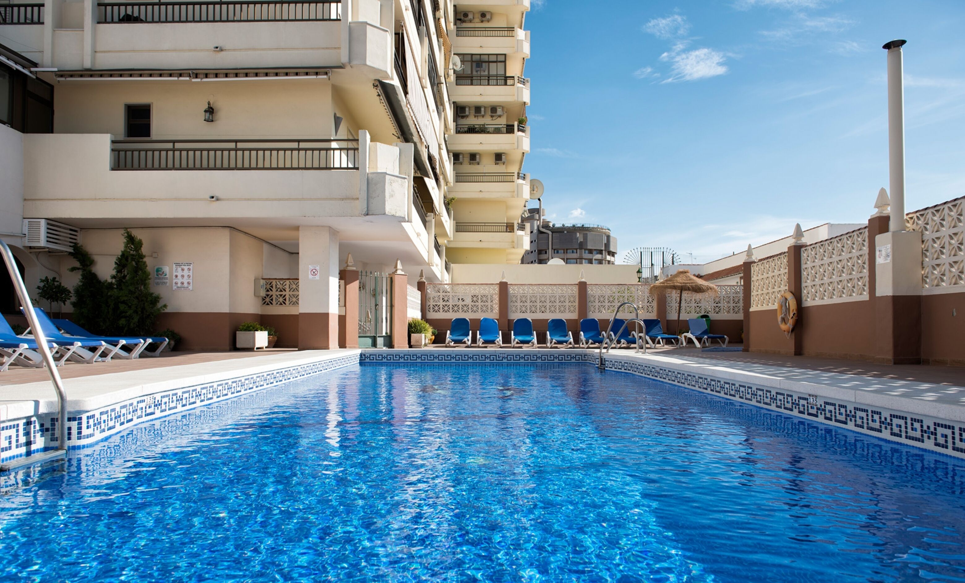 A swimming pool with blue tiles, surrounded by lounge chairs and a building with balconies.