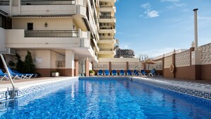 Une piscine aux carreaux bleus, entourée de chaises longues et bordée d’un bâtiment doté de balcons.