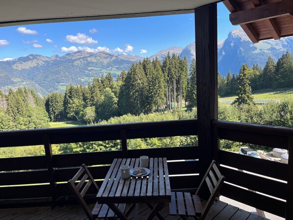 A wooden deck with a table and chairs, overlooking a mountainous landscape with trees and a clear sky.