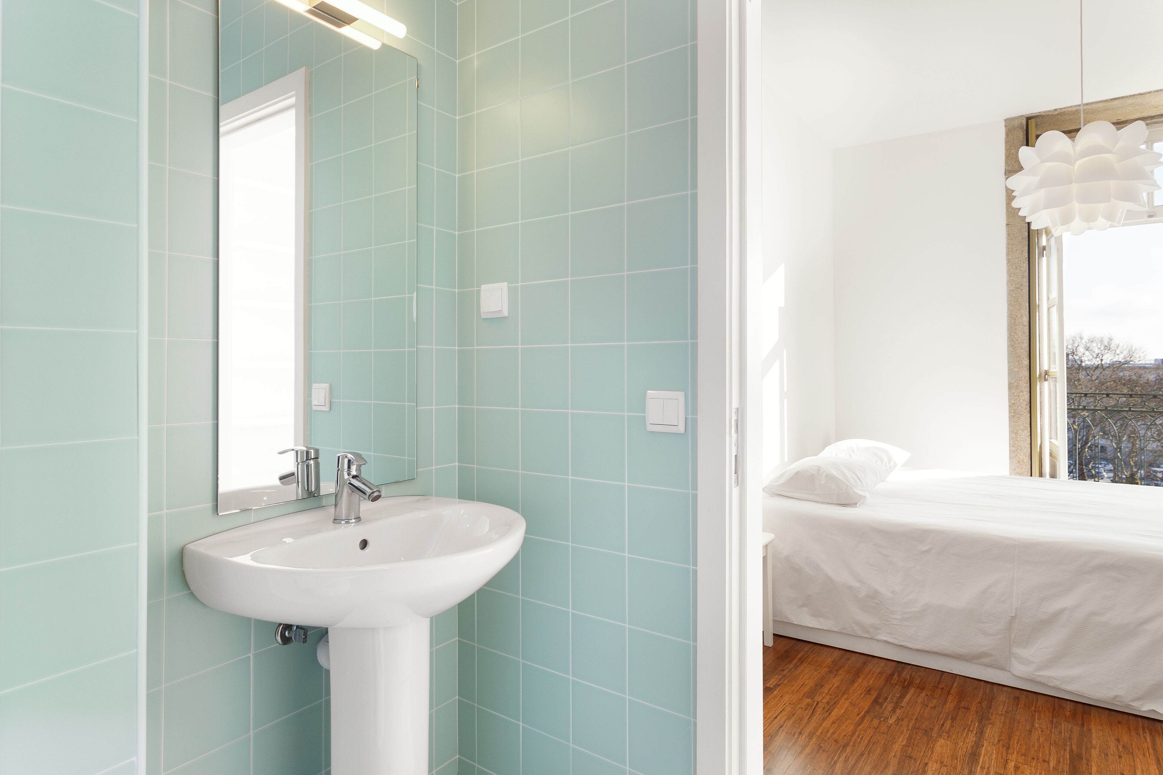 A modern bathroom with a white sink, a mirror, and a view of a bedroom with a bed and a window.