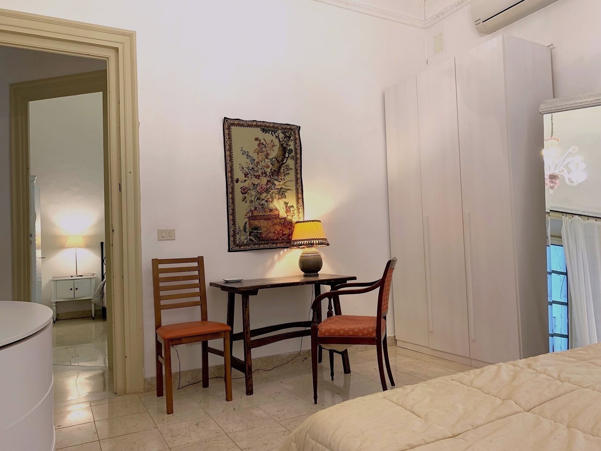 A hotel room with a bed, two wooden chairs, a small table with a lamp, and a painting on the wall.