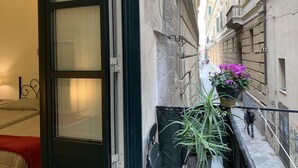 A narrow urban street with a building on one side and a balcony with a potted plant on the other.