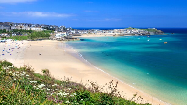 A coastal beach with clear turquoise waters, a sandy shore, and a town in the background.
