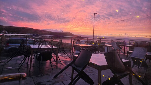 An outdoor seating area with tables and chairs, overlooking a body of water and a sunset.