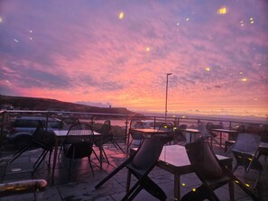 An outdoor seating area with tables and chairs, overlooking a body of water and a sunset. - Spacious 3 Bed Apartment | Spectacular Sea Views | Close to Town & Beach (Bude)