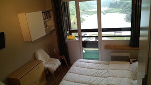 Un dormitorio con cama, una silla, un escritorio pequeño, una estantería con libros y una ventana con vistas a un paisaje verde.