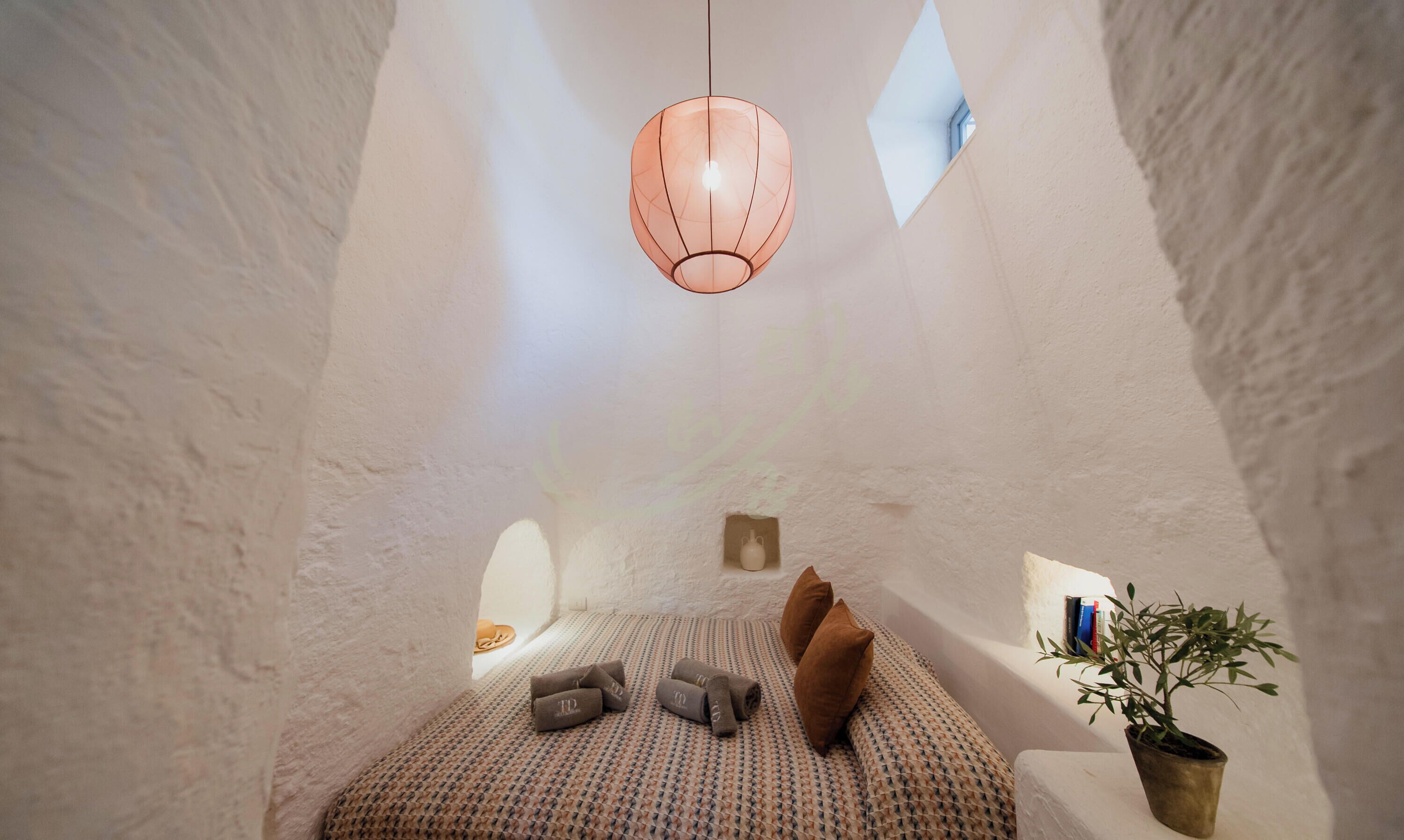 Ein gemütliches Zimmer mit Bett, Kissen, einer Pendelleuchte, einem kleinen Bücherregal und einem Blumentopf.