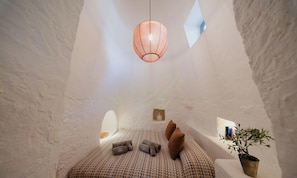 Una habitación acogedora con cama, almohadas, una lámpara colgante, un pequeño estante con libros y una planta en maceta.