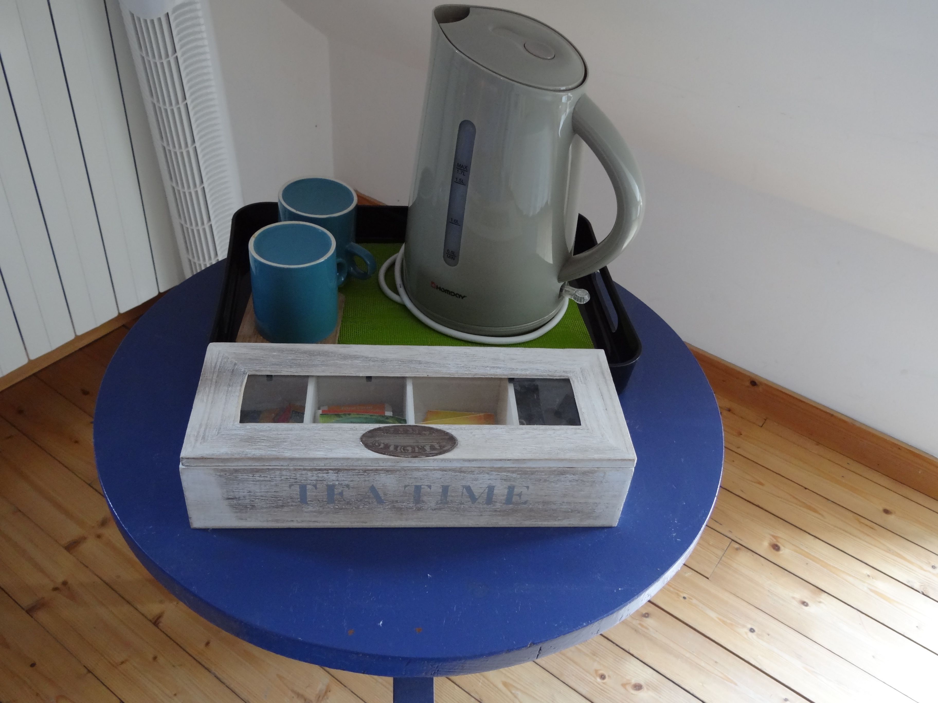 Una pequeña mesa con un tablero azul, una caja blanca con la etiqueta 'TEA TIME', una tetera eléctrica gris y dos tazas azules.