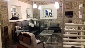 A cozy living room with a stone wall, a black leather sofa, a glass coffee table, and a white chair. - charming country cottage for rent  (Ave et Auffe)