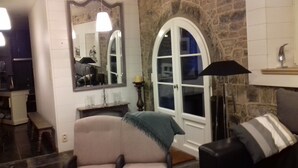 A cozy living room with a stone wall, a large arched window, a fireplace, and a comfortable armchair. - charming country cottage for rent  (Ave et Auffe)
