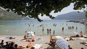 Ein voller Strand mit badenden und sonnenden Menschen, umgeben von Bergen und wolkenlosem Himmel.