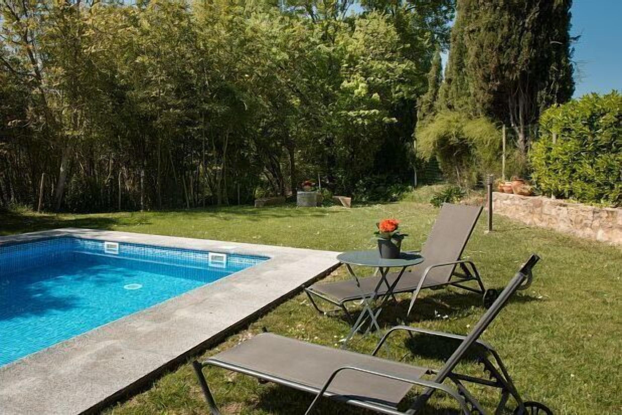A swimming pool with a tiled edge, surrounded by grass and trees. There are two lounge chairs and a small table with a flower pot.