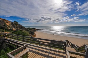 A wooden boardwalk leading to a beach with a rocky cliff on one side and a clear sky above.