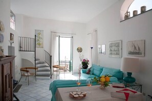 Un salón luminoso con un sofá azul aguamarina, una mesa de comedor con bebidas y pasteles, una escalera y un balcón con vistas.