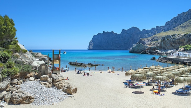 A beach with clear blue water, rocky terrain, and umbrellas. - VILLA ALMADRAVA - VACATION PARADISE (Pollensa)