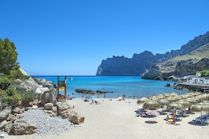 Een strand met helderblauw water, rotsachtig terrein en parasols.