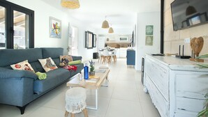 A modern living room with a blue sofa, a wooden coffee table, and a white round ottoman. There is a kitchen in the background with a dining area. - VILLA ALMADRAVA - VACATION PARADISE (Pollensa)