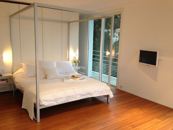 A modern bedroom with a four-poster bed, a bedside table with a lamp, and a view of the outdoors through large windows.