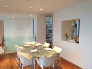 Un espace repas moderne avec une table en verre, des chaises blanches et une vue sur un espace extérieur rocheux, visible à travers une fenêtre.