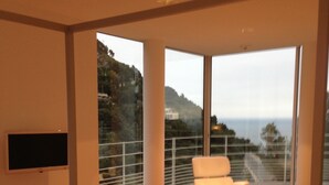 A modern bedroom with a large window, a flat-screen TV, a bed with a tray of food, and a white chair. - Spectacular Panoramic Views of Cap Ferrat & Beaulieu. (Villefranche-Sur-Mer)