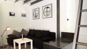 Ein modernes Wohnzimmer mit einem schwarzen Sofa, einem weißen Couchtisch und einer Stehlampe. An der Wand befinden sich zwei gerahmte Bilder, und eine Treppe führt auf eine andere Ebene.