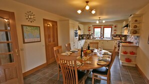 A dining area with a wooden table set for four, surrounded by chairs. A kitchen with white cabinets and red accents is visible in the background.
