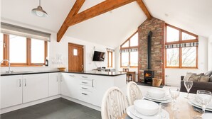 A modern kitchen with white cabinetry, a dining area with a wooden table set for six, and a stone fireplace. - Combermere Cottage - Two Bedroom Cottage, Sleeps 4 (Bude)