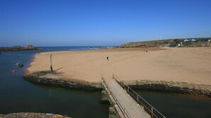 A coastal beach with a pier extending into calm waters, a lighthouse in the distance, and a few scattered buildings. - Combermere Cottage - Two Bedroom Cottage, Sleeps 4 (Bude)