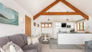 A modern living room with a grey sofa, a dining area with a white table and chairs, a kitchen with white cabinets and a wooden ceiling, and a large window providing natural light. - Combermere Cottage - Two Bedroom Cottage, Sleeps 4 (Bude)