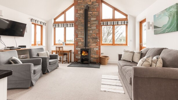 A living room with a stone fireplace, grey sofas, and a television. - Combermere Cottage - Two Bedroom Cottage, Sleeps 4 (Bude)