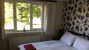 A hotel room with a large bed, two red towels, a radiator, a window with curtains, and a floral wallpaper pattern. - Well Equipped Family Holiday Home With Garden In A Quiet rural location. (Sutton)