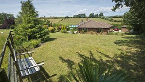 A grassy area with a wooden bench, a brick house, and a car parked in the background. - Well Equipped Family Holiday Home With Garden In A Quiet rural location. (Sutton)