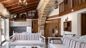 Interior rústic amb paret de pedra, bigues de fusta, cuina i llit amb roba de llit estriada.