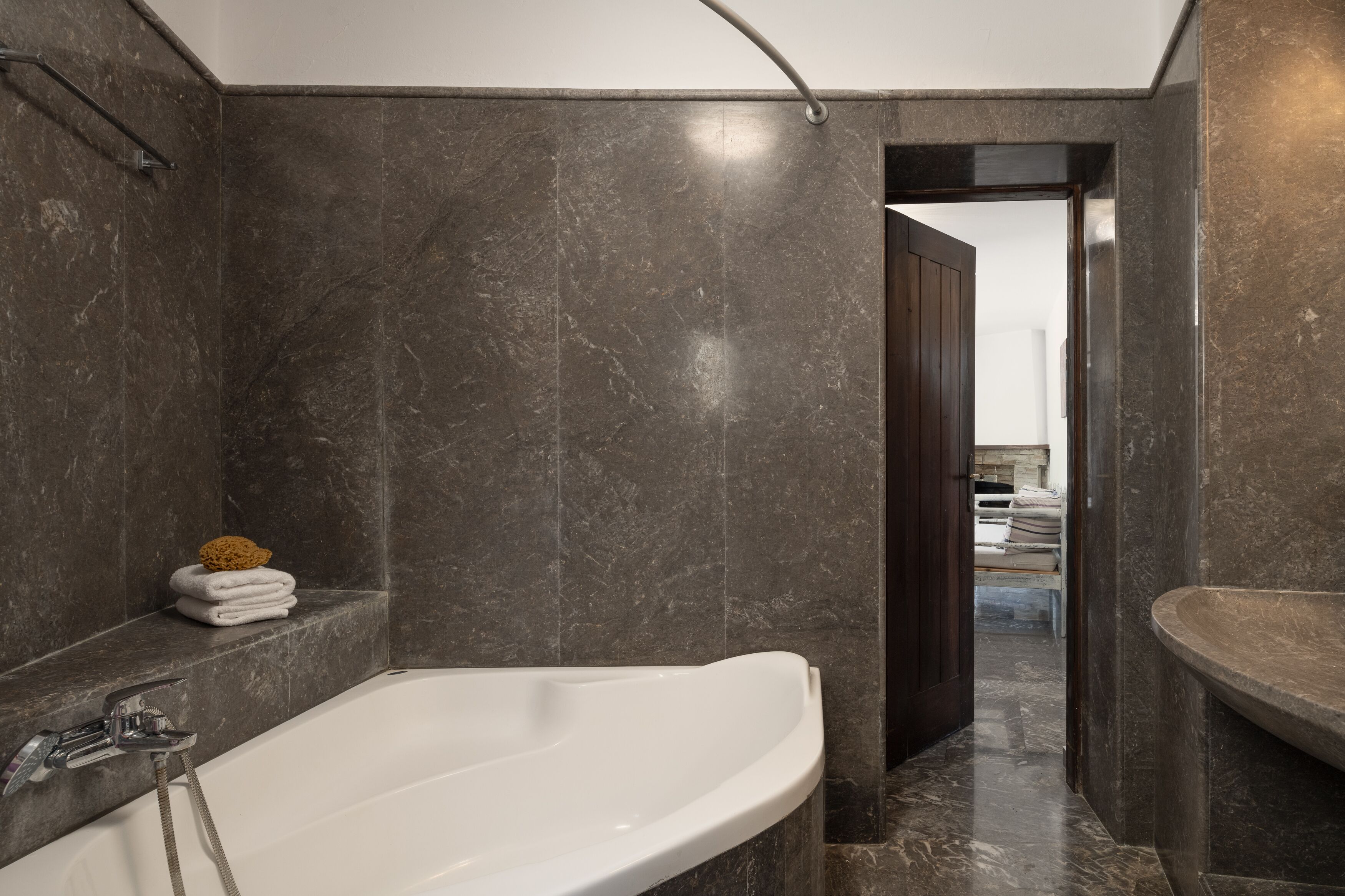 A modern bathroom with a large bathtub, dark marble walls, and a visible doorway leading to another room.
