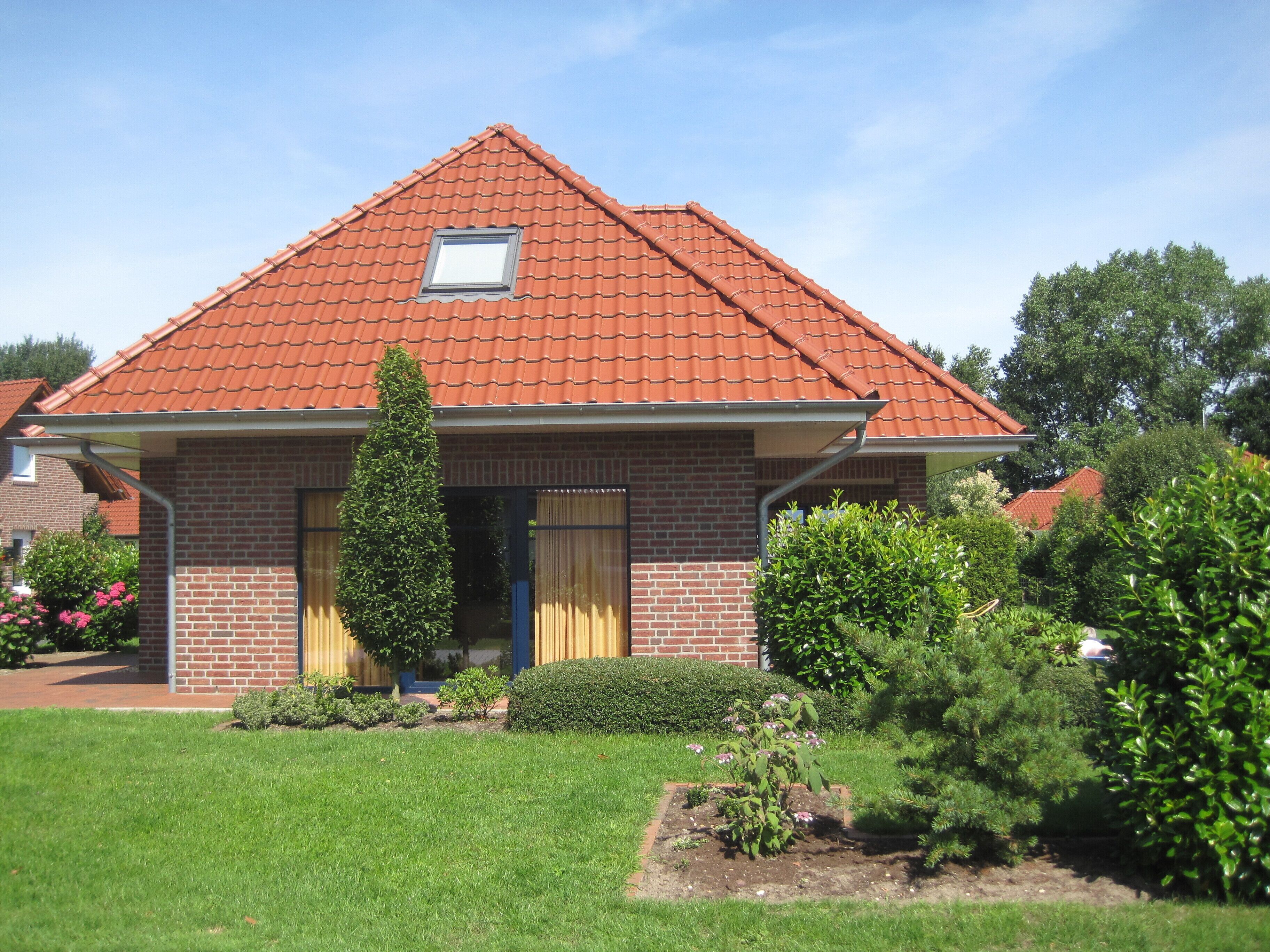 A two-story brick house with a red-tiled roof, a small window, and a well-maintained garden with various plants and flowers.