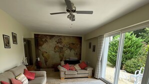 Una sala de estar con un sofá color beige, un sofá más pequeño con cojines rojos, un tapete gris, un ventilador de techo y un ventanal con vistas a la vegetación.