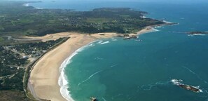 Vista aérea de una zona costera con una playa de arena, un mar azul cristalino y un paisaje montañoso al fondo.