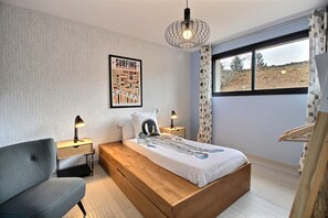 Un dormitorio con cama, una silla, una mesita de noche, una lámpara, una ventana con vistas y un cartel de surf en la pared.