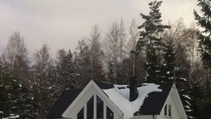 Uma casa branca de dois andares com telhado íngreme, rodeada por terreno e árvores cobertos de neve.