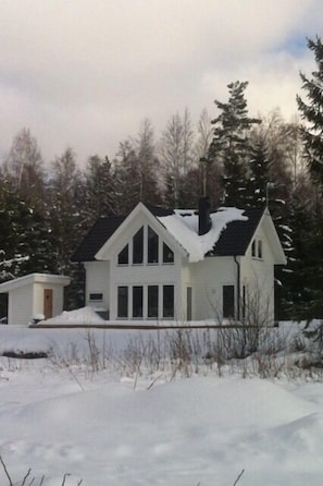 A white two-story house with a steeply pitched roof, surrounded by snow-covered ground and trees.