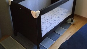 Berço infantil com lateral de tela e base sólida, posicionado sobre um piso de madeira com tapete azul.