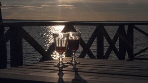 Dois copos sobre um deck de madeira com guarda-corpo, com vista para um corpo d'água onde a luz do sol se reflete.