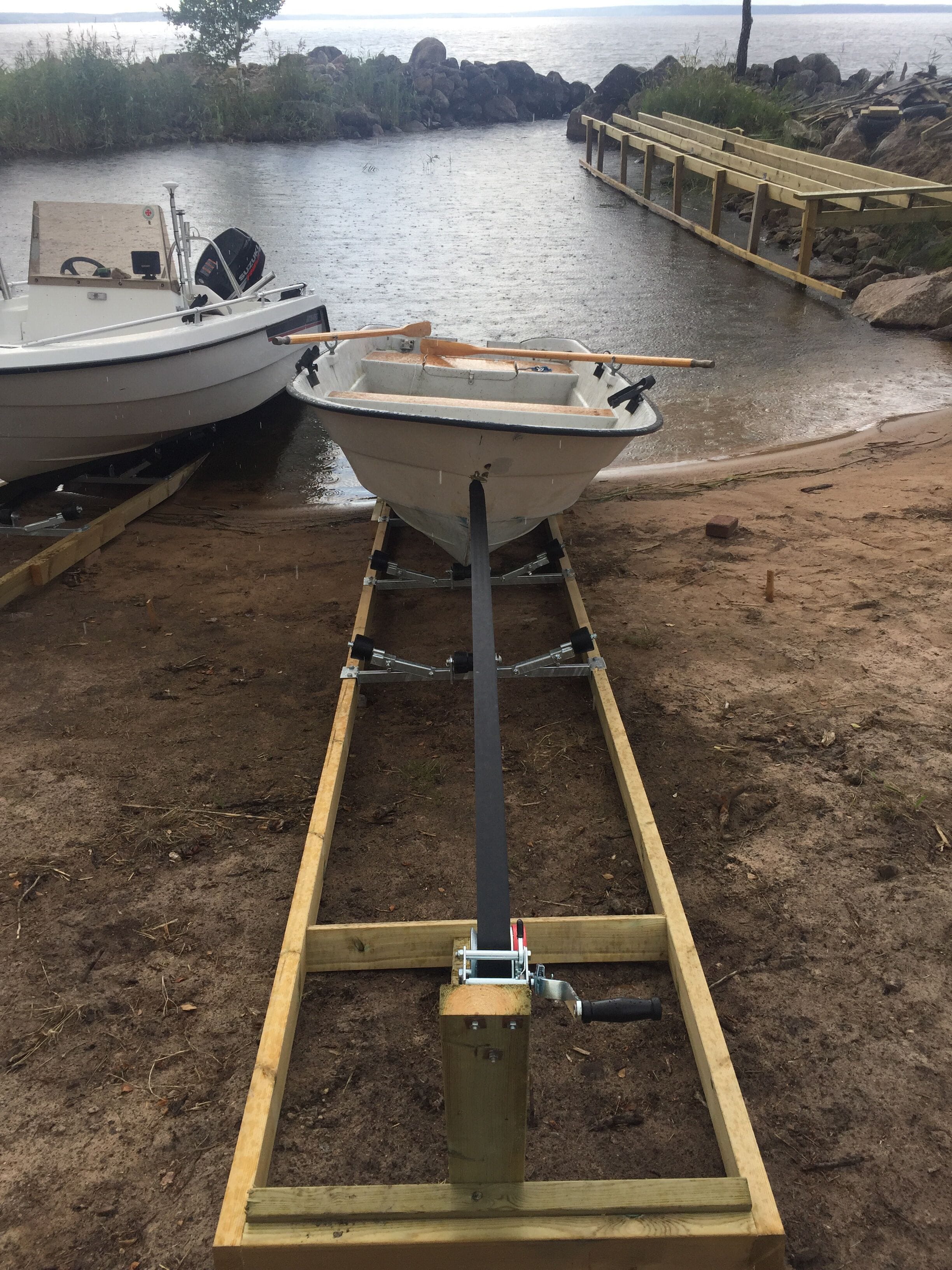 Ein Boot auf einem Anhänger an einem sandigen Strand mit felsiger Küstenlinie und einem Holzsteg im Hintergrund.