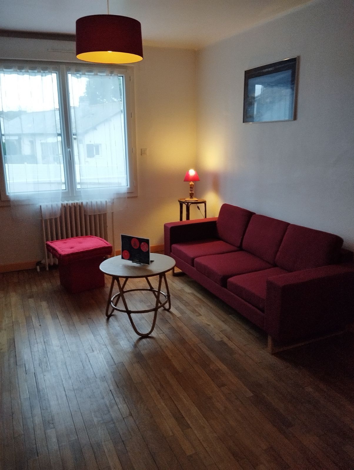 A living room with a red sofa, a small round table, a red lamp, and a window with a view of buildings.