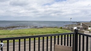 A coastal view from a balcony with a railing, a grassy area, and a building with a chimney. - Northern Hideaways Seascape (Seahouses)