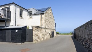 Una calle residencial con una casa blanca, un muro de piedra y una vista despejada del mar.